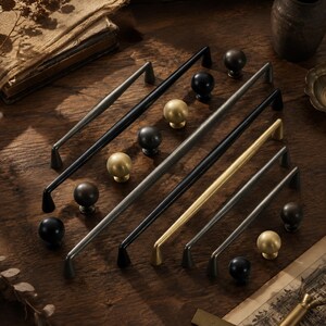 May include: A collection of cabinet hardware including several long, slender metal drawer pulls and round knobs. The hardware features a variety of finishes, including matte black, brushed gold, and dark metallic grey, arranged in a scattered layout.