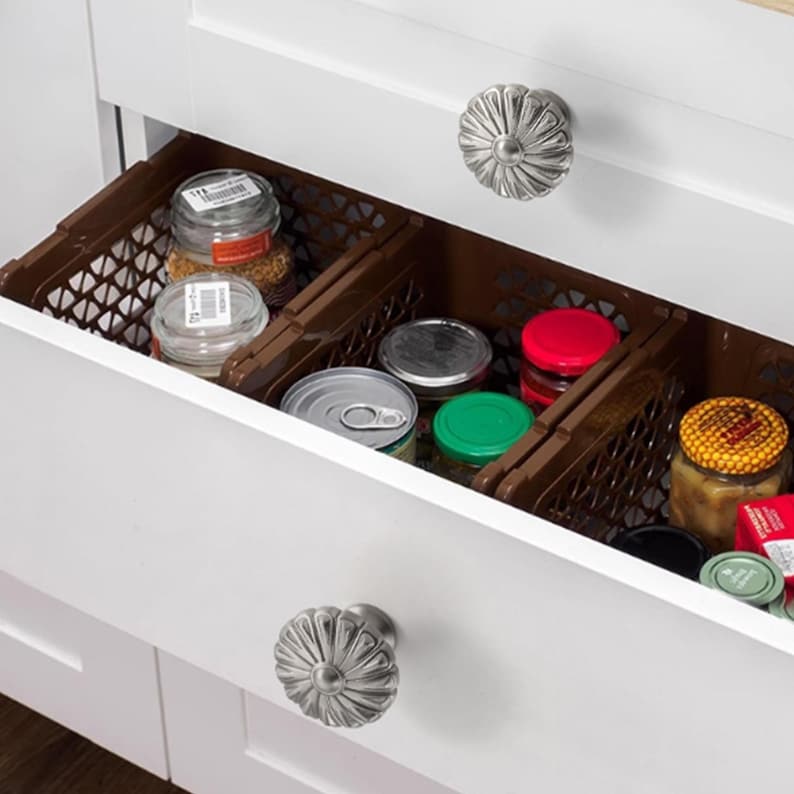 Op de afbeelding: Een open witte keukenlade met twee zilveren bloemvormige knoppen. Binnenin bevinden zich bruine plastic opbergkratten met diverse glazen potten met metalen deksels, een metalen blik en een pot met een geel deksel met honingraatpatroon.