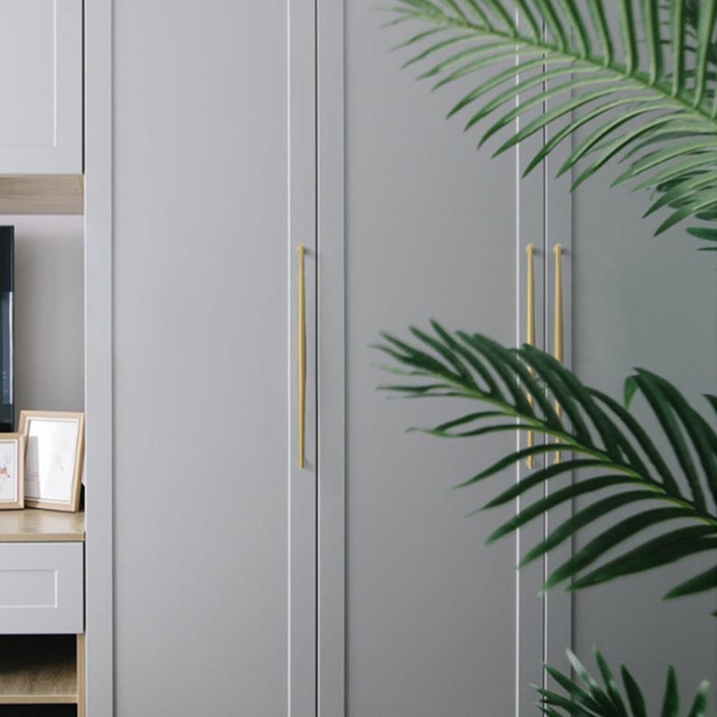 May include: A set of tall, slender, brushed gold cabinet pulls mounted vertically on light gray, flat-panel double doors. The hardware features a smooth, elongated cylindrical design that contrasts with the matte finish of the cabinetry.