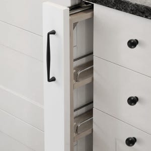 May include: A narrow, white pull-out kitchen cabinet organizer with a black handle. The unit features light wood shelves with thin, horizontal silver metal rails to secure items. It is positioned between white cabinets with black round knobs.