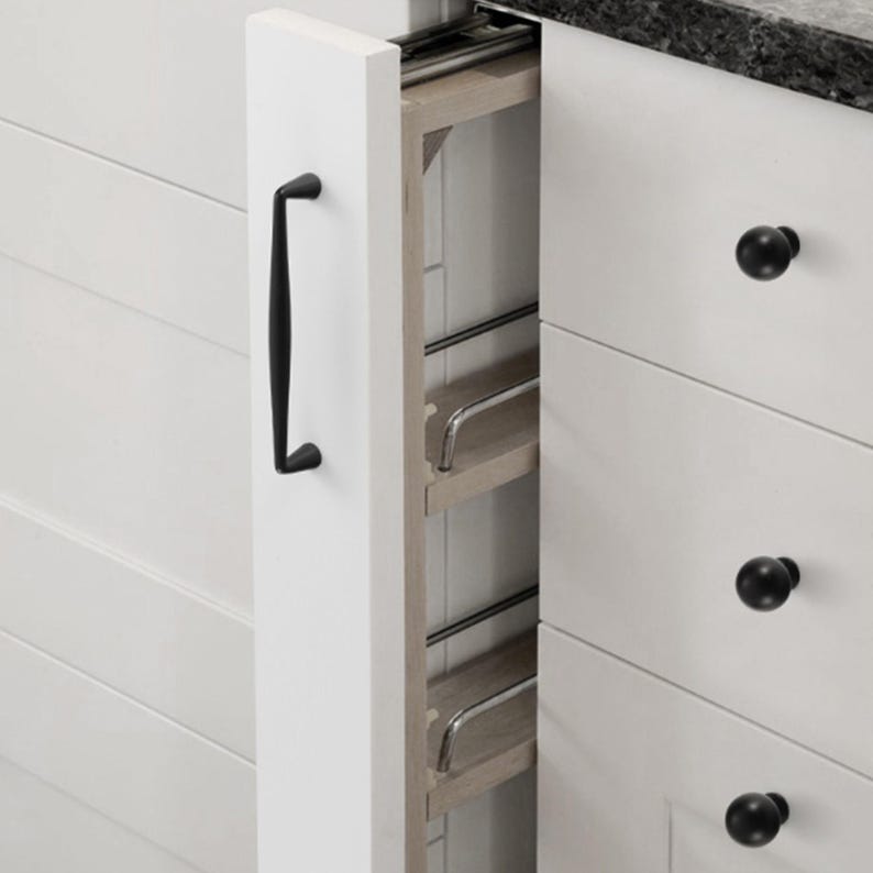 May include: A narrow, white pull-out kitchen cabinet organizer with a black handle. The unit features light wood shelves with thin, horizontal silver metal rails to secure items. It is positioned between white cabinets with black round knobs.