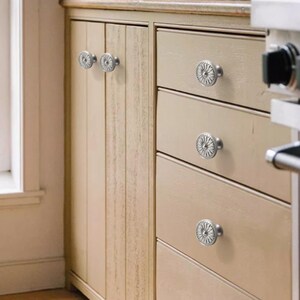 Op de afbeelding: Een set ronde, zilverkleurige metalen kastknoppen met een gedetailleerd, stralend zonnestraal- of bloemmotief. Deze ronde knoppen zijn gemonteerd op lichtbeige houten keukenkasten, inclusief zowel verticale kastdeuren als horizontale lades.