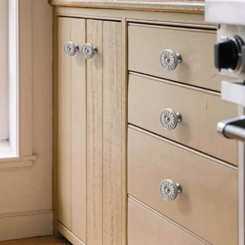 Op de afbeelding: Een set ronde, zilverkleurige metalen kastknoppen met een gedetailleerd, stralend zonnestraal- of bloemmotief. Deze ronde knoppen zijn gemonteerd op lichtbeige houten keukenkasten, inclusief zowel verticale kastdeuren als horizontale lades.