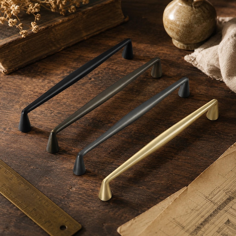 May include: Four metal cabinet pulls arranged diagonally, featuring a matte black, dark bronze, charcoal gray, and brushed gold finish. Each handle has a slender, slightly arched bar with conical bases. A brass ruler is visible in the corner.