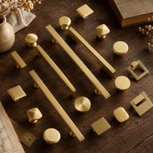 Peut inclure: Une collection de quincaillerie d'armoire en or brossé comprenant diverses poignées de tiroir rectangulaires longues, des boutons ronds, des boutons carrés et des poignées tombantes carrées disposées en vrac. Finition métallique mate.