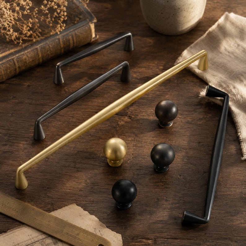 May include: A collection of cabinet hardware including three long metal bar pulls in matte black and brushed gold finishes, alongside four spherical knobs in matching black and gold tones, arranged with a wooden ruler for scale reference.