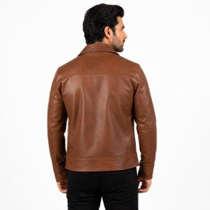 Puede incluir: Una chaqueta de cuero color canela con cuello cl&aacute;sico, mangas largas con pu&ntilde;os abotonados y dobladillo recto. La chaqueta se muestra desde atr&aacute;s, exhibiendo una costura horizontal en la parte superior y costuras verticales en los lados.