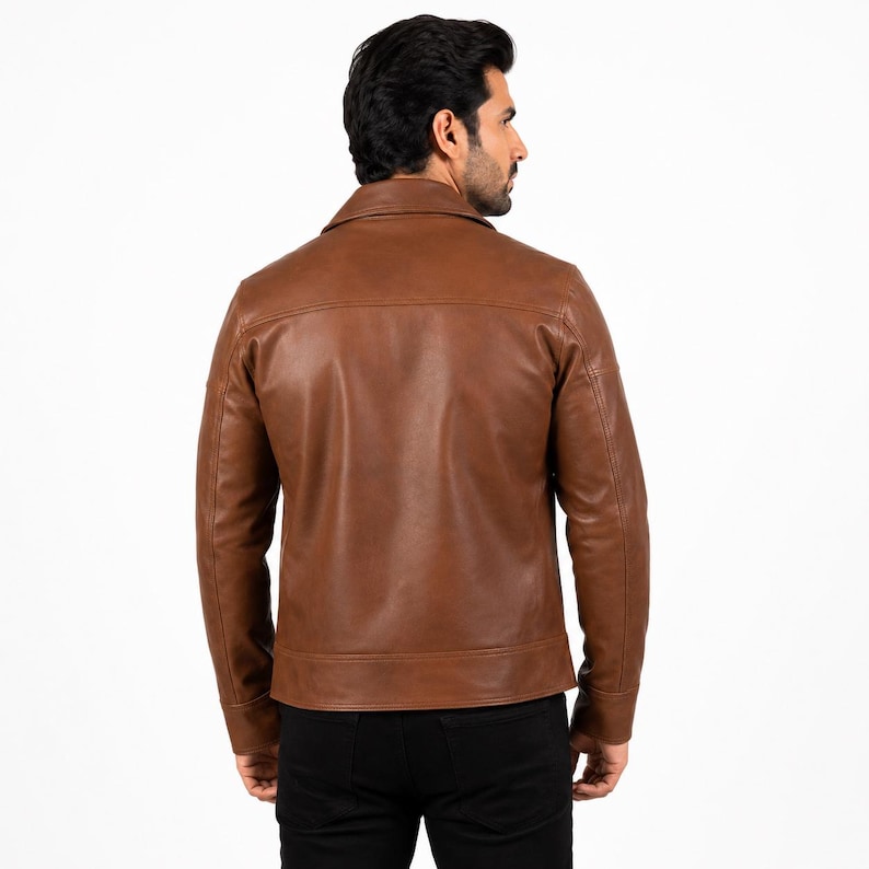 Puede incluir: Una chaqueta de cuero color canela con cuello cl&aacute;sico, mangas largas con pu&ntilde;os abotonados y dobladillo recto. La chaqueta se muestra desde atr&aacute;s, exhibiendo una costura horizontal en la parte superior y costuras verticales en los lados.