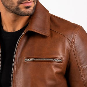 Puede incluir: Una chaqueta de cuero marr&oacute;n con cuello cl&aacute;sico, una cremallera frontal de metal plateado de longitud completa y un bolsillo horizontal con cremallera en el lado izquierdo del pecho. La chaqueta se lleva sobre una camiseta negra lisa.