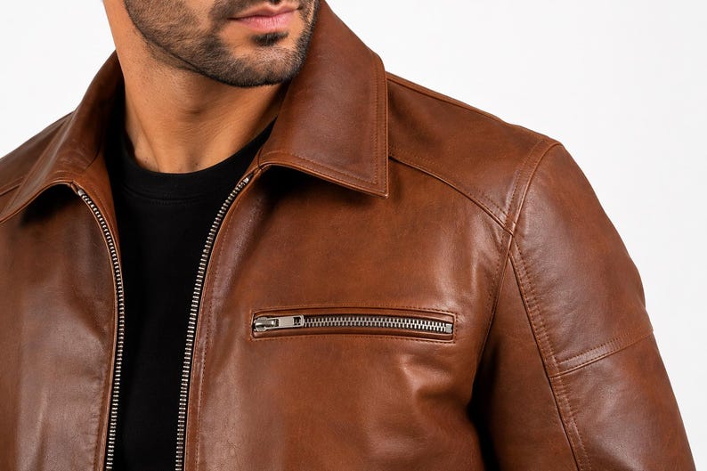 Puede incluir: Una chaqueta de cuero marr&oacute;n con cuello cl&aacute;sico, una cremallera frontal de metal plateado de longitud completa y un bolsillo horizontal con cremallera en el lado izquierdo del pecho. La chaqueta se lleva sobre una camiseta negra lisa.