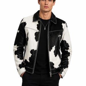 Puede incluir: Una chaqueta con estampado de vaca en blanco y negro, con cuello vuelto, cremallera frontal completa y dos bolsillos con cremallera en el pecho. La prenda tiene mangas largas con puños y costuras blancas visibles en todo el diseño.