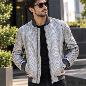 Puede incluir: Una chaqueta bomber plateada con un patrón texturizado de piel de serpiente, que presenta un cuello, puños y dobladillo acanalados en gris oscuro. La chaqueta tiene un cierre de cremallera frontal completo y dos bolsillos laterales, sobre una camiseta negra.