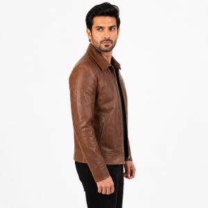 Puede incluir: Una chaqueta de cuero marr&oacute;n con cuello cl&aacute;sico, cremallera frontal completa y dos bolsillos laterales con cremallera. La chaqueta tiene mangas largas con pu&ntilde;os y se lleva sobre una camiseta negra y pantalones negros, con un acabado liso.