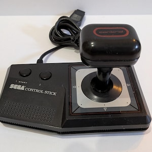 May include: A black Sega Control Stick video game controller featuring a joystick with a rectangular top, two circular buttons, and a wired connection. The base displays the Sega logo and the text Control Stick, with a silver square plate surrounding the joystick base.