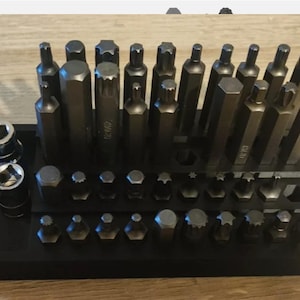 May include: A comprehensive set of dark grey metal tool bits and two cylindrical sockets arranged in a black rectangular organiser tray. The collection includes various shapes such as star, hexagonal, and splined heads, all neatly organised in rows.