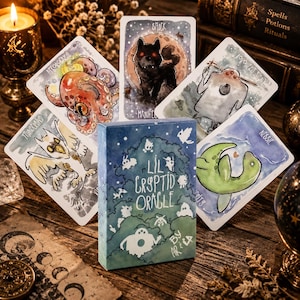 May include: A deck of Lil Cryptid Oracle cards featuring watercolor illustrations of mythical creatures including an octopus, a wolf, a ghost, a thunderbird, and Nessie. The box displays the title Lil Cryptid Oracle by Arila in white text.