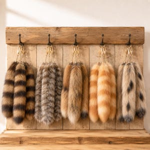 May include: Five decorative faux fur tail keychains hanging from metal hooks. The tails feature various patterns including brown and black stripes, grey mottled fur, solid tan, orange and white stripes, and tan with black spots, all with gold clips.