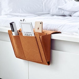 May include: A tan felt bedside storage caddy featuring multiple pockets, holding a white remote control, a book titled WEEKEND IN PARIS, and a gold smartphone. The organiser is draped over the edge of a white bed frame.