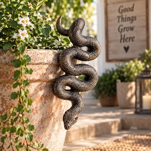 May include: A dark bronze-coloured snake sculpture with a textured, scale-like pattern draped over the rim of a large, weathered terracotta planter. The snake is coiled in an S-shape, with its head pointing downwards towards the base of the pot.