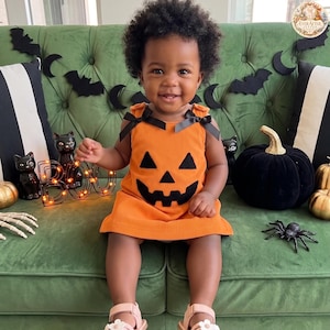 Peut inclure: Une robe orange sans manches avec un motif de visage de citrouille d'Halloween noir sur le devant et des liens en ruban noir aux épaules. La robe est confectionnée dans un tissu côtelé et présente une silhouette droite et simple.