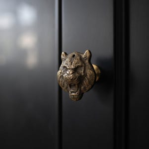 May include: A brass-toned metal cabinet knob shaped like a tiger head with a detailed mane, pointed ears, and an open mouth. The hardware is mounted onto a smooth, matte black surface, featuring a small circular hole at the top of the tiger's head.