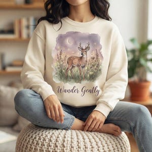 Puede incluir: Una sudadera de cuello redondo color crema con un gráfico estilo acuarela de un ciervo de pie en un campo de flores silvestres bajo una luna creciente. El texto "Wander Gently" está impreso en una fuente cursiva oscura debajo.