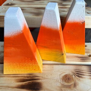 May include: Three triangular wooden blocks painted to resemble candy corn. Each block features a horizontal color gradient starting with white at the top, transitioning to orange in the middle, and ending with a bright yellow base.
