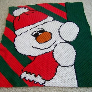 May include: A crocheted snowman blanket with a red and green striped background. The snowman is wearing a red hat and scarf.