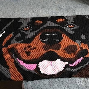 Puede incluir: Manta de crochet con un primer plano de la cara de un Rottweiler. El pelaje del perro es negro, marrón y gris, y su lengua es rosa. La manta está hecha con hilo blanco y negro.