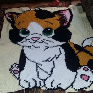 May include: A crocheted blanket featuring a cartoon cat with black, orange, and white fur. The cat has green eyes and a pink nose. The blanket is made with a white yarn and has a textured surface.