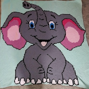 May include: A crocheted baby blanket featuring a grey elephant with pink ears and a light blue background.