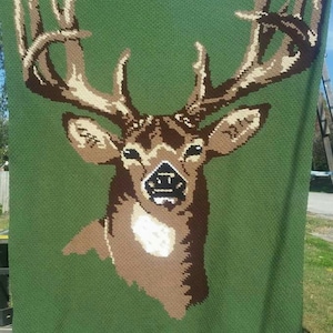 Può includere: Una coperta di uncinetto verde con un disegno di testa di cervo marrone e bianco. La coperta ha il testo "crocheted by tester Catina" in basso.