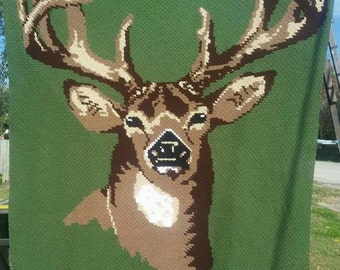 May include: A green crocheted blanket with a brown and white deer head design. The blanket has the text "crocheted by tester Catina" at the bottom.