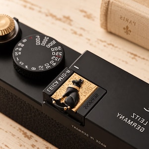 Puede incluir: Una placa superior de cámara negra con un dial de velocidad de obturación circular con números blancos, una perilla de cuerda dorada y una incrustación decorativa dorada que representa un gato negro con grabados jeroglíficos.