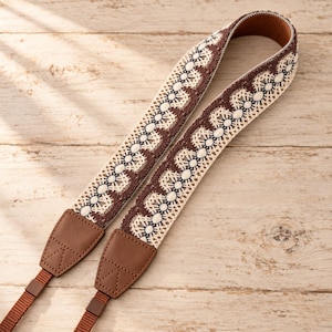 May include: A camera strap featuring a woven fabric band with a repeating brown and cream lace-like pattern. The strap includes brown faux leather end tabs with triangular stitching and thin brown nylon webbing connectors at the bottom ends.