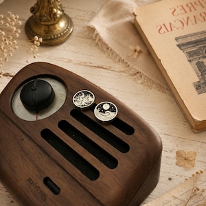 May include: A dark brown wooden radio with a black circular dial and three horizontal speaker slots. Two small circular pins featuring black and white illustrations of a mountain landscape and a moonlit ocean wave rest on the radio surface.
