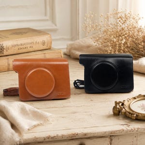 Pode incluir: Duas bolsas para câmara, uma em castanho claro e outra em preto, ambas com um acabamento liso e estruturado semelhante a couro, com uma secção circular para a lente e um pequeno logótipo em relevo no canto inferior de cada capa.