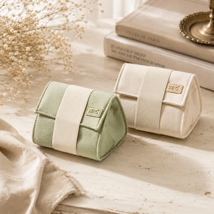 Puede incluir: Dos pequeñas bolsas de tela rectangulares con perfil triangular, una en verde salvia y otra en crema. Cada bolsa presenta una banda vertical central y un pequeño parche rectangular en la solapa con el texto XBYS impreso en tinta negra.