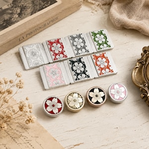 Puede incluir: Una colección de botones metálicos rectangulares y circulares con motivos de flores de cerezo. Los botones rectangulares son blancos, rojos, grises, verdes, rosas, negros y naranjas. Los botones circulares tienen fondos rojo, dorado, negro y rosa.