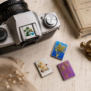 Puede incluir: Una cámara vintage plateada con un inserto cuadrado de una isla tropical, acompañada de tres pines rectangulares con un diseño de tigre azul, un motivo floral plateado y un patrón de ramas moradas con detalles dorados.