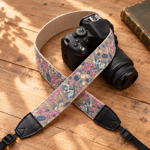 Op de afbeelding: Een camerariem met een beige achtergrond en een herhalend patroon van blauwe vlinders, roze bloemen en donkerblauwe bladeren. De riem heeft zwarte lederen uiteinden en verstelbare zwarte nylon banden bevestigd aan een zwarte digitale camera.