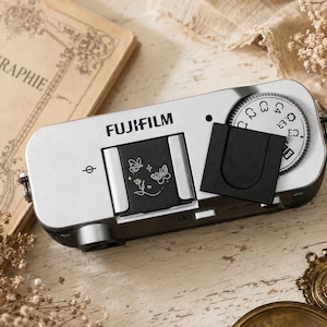 May include: A silver Fujifilm digital camera top view featuring a black hot shoe cover engraved with a white line art design of two butterflies and a flower. The camera includes a circular mode dial with alphanumeric settings and a black shutter button.