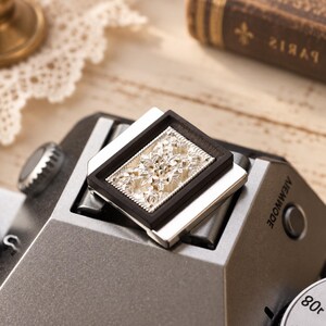 May include: A rectangular camera hot shoe cover featuring a silver-toned metal plate with an intricate, embossed floral pattern framed by a dark, smooth border. The accessory is mounted on the top of a grey camera body with visible text.