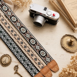 May include: Two camera straps featuring intricate woven patterns. One strap displays a black background with light blue floral motifs, while the other features a geometric diamond design in shades of black, white, and tan. Both have tan leather ends.