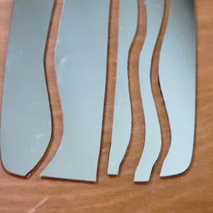 May include: A set of four silver-colored metallic pieces with a reflective finish. Each piece features a distinct shape, with some having straight edges and others having curved, wave-like contours, arranged side by side in a vertical orientation.