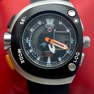 May include: A Citizen Eco-Drive diver's watch with a black rubber strap and a silver-tone case. The black dial features analog hands with orange accents, a digital display, and text including SEL, MODE, LOG, DIVE, and MAX. DEPT. ft. The watch has a rotating bezel.