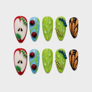 May include: A set of ten press-on nails featuring nature-themed designs including a sliced apple with a green caterpillar, blue sky with red ladybugs, green leaves with water droplets, green leaves with caterpillars, and orange butterfly wings.