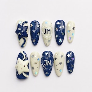 May include: A set of ten press-on nails featuring a celestial theme with deep navy blue and cream white bases. The designs include holographic star sequins, a raised dark blue star charm with a cat face, a cream star charm, and heart-shaped accents with the letters JM and JN.