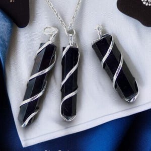 May include: Three black gemstone pendants wrapped in silver wire, hanging from silver chains. The pendants are displayed on a white surface with a blue background.