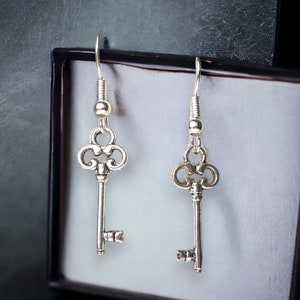 May include: A pair of silver key-shaped earrings with intricate details. The earrings are hanging from a black box.